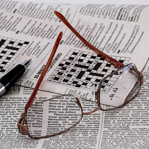 Na zdjęciu widać okulary, leżące na rozłożonej gazecie z tekstem i krzyżówką. Po lewej stronie jest widoczny fragment długopisu.