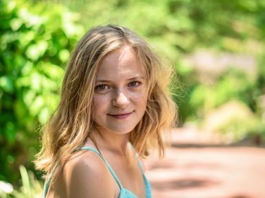 Młoda blondynka uśmiecha się delikatnie na tle zieleniących się drzew.