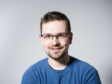 Krótkowłosy szatyn w okularach i błękitnym swetrze uśmiecha się, pozując na szarym tle.