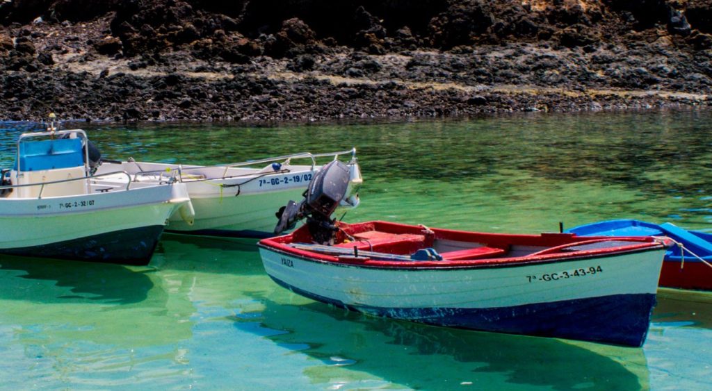 Plakat informujący o spotkaniu podróżniczym z Markiem Dąbrowskim. Na plakacie zdjęcie wyspy Fuerteventura - plaży z kolorowymi łodziami