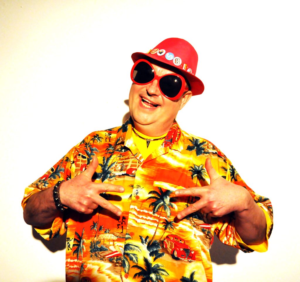 Na zdjęciu uśmiechnięty mężczyzna w ciemnych okularach, czerwonym kapeluszu i kolorowym stroju.