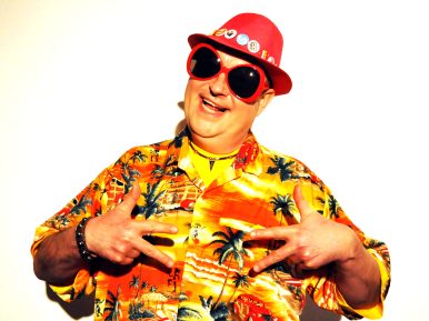 Na zdjęciu uśmiechnięty mężczyzna w ciemnych okularach, czerwonym kapeluszu i kolorowym stroju.